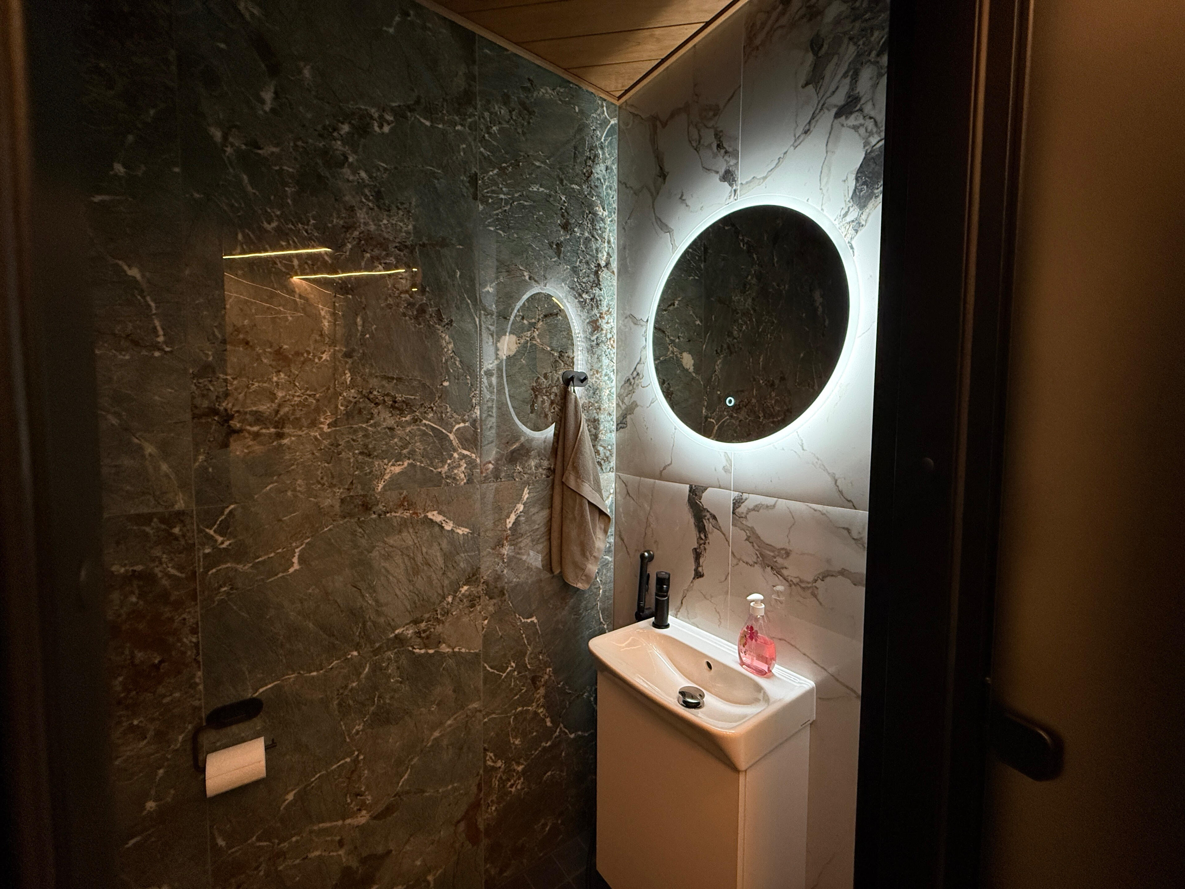 Vaaralan Saunan tyylikäs wc-tila Vaaralan Saunan moderni wc-tila, jossa on marmorikuvioidut seinät, pesuallas ja valaistu pyöreä peili