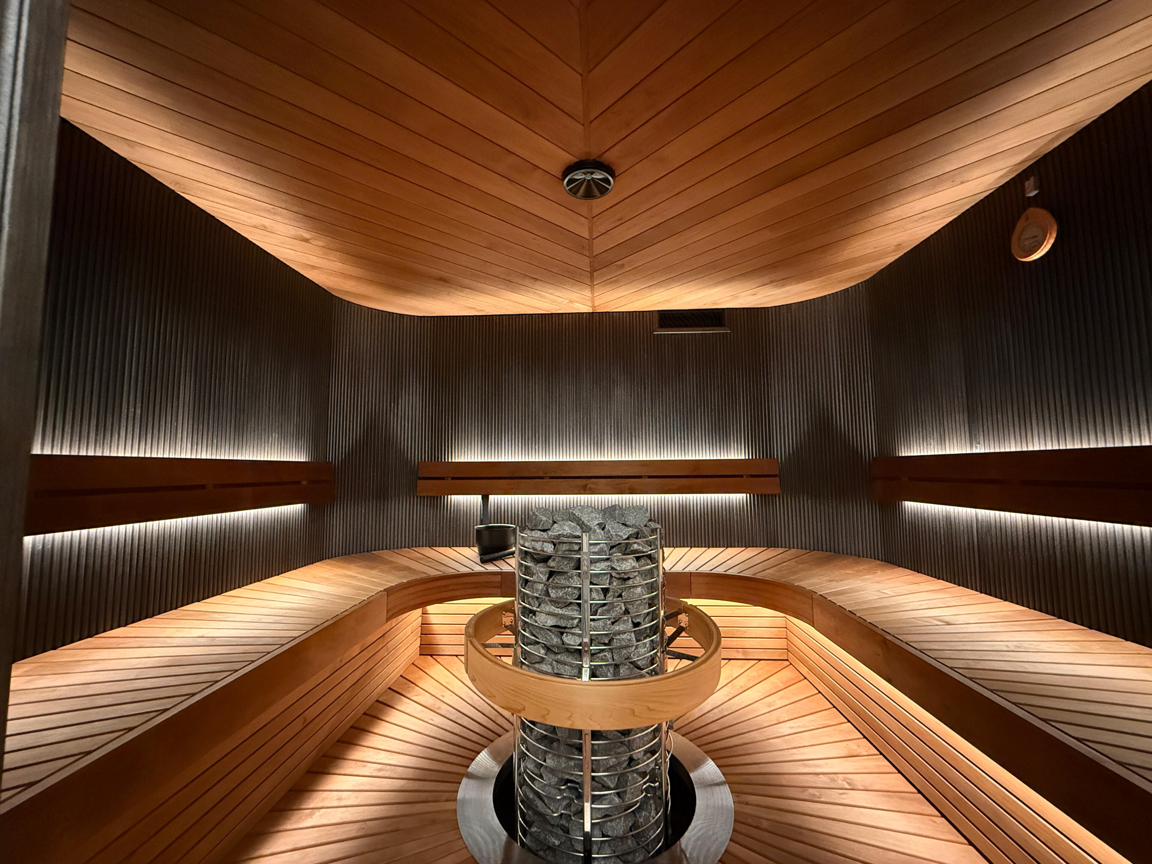 Vaaralan Sauna logo Vaaralan Saunan moderni sauna, jossa on suuri kivikiuas, kaarevat puulauteet ja tunnelmallinen epäsuora valaistus